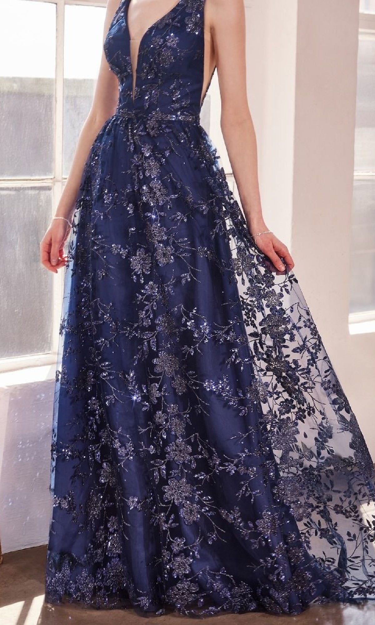 A woman wearing the Amoris T606 Embroidered-Lace Long Prom Dress—a sleeveless, deep V-neck navy blue gown with floral lace and sequin details—stands indoors by a window.